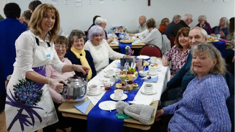A table of people smiling with one person standing up holding a teapot, looking at the camera